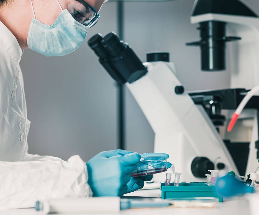 Prillide ja maskiga laborant uurib käes olevat analüüsi ja taustal on laual mikroskoop