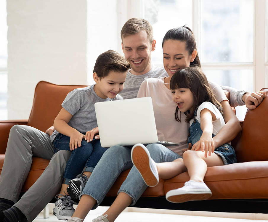 Neljaliikmeline perekond istub pruunil diivanil ja vaatavad naerdes sülearvutit