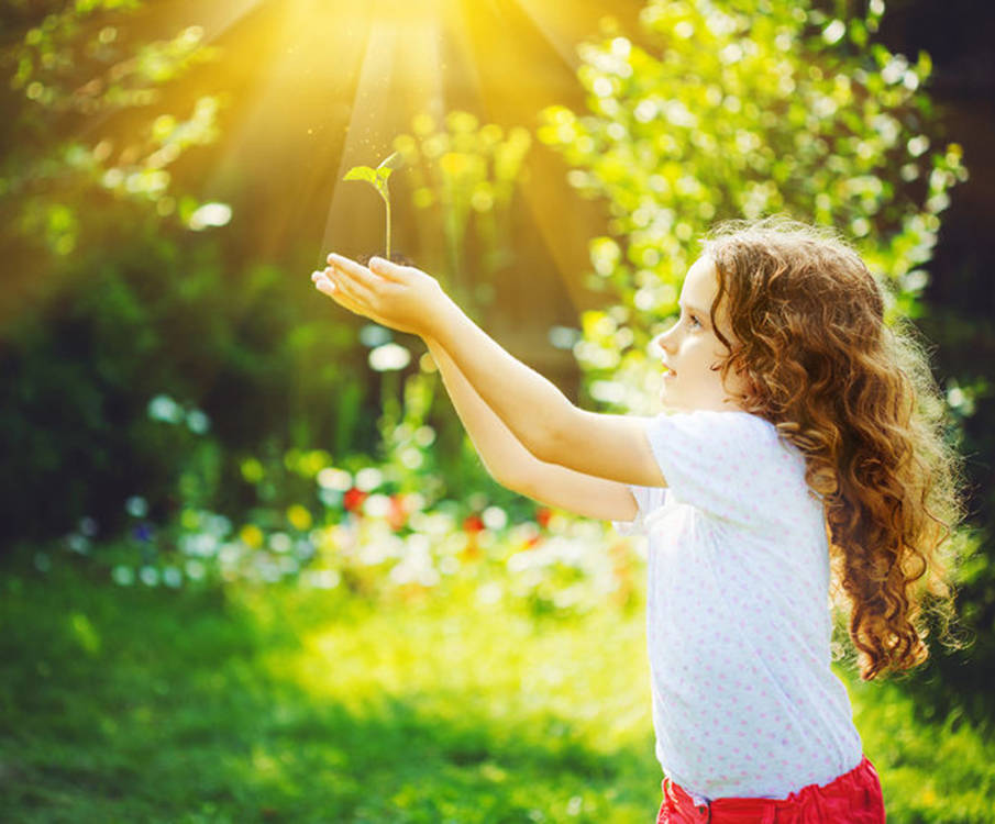 The girl stands outdoors and holds a plant in the palm of her hand in the sun